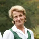 Linde Rainer mit kurzen, blonden Haaren trägt weiße Bluse und grünes, ärmelloses Kleid vor unscharfem Hintergrund.