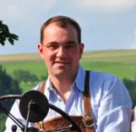 Andreas Hager trägt hellblaues Hemd und braune Lederhosenträger, sitzt vor grünem Feld und blauem Himmel.