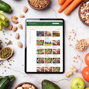 Tablet mit Gutes vom Bauernhof Website, umgeben von frischem Gemüse, Hülsenfrüchten und Nüssen auf einer hellen Oberfläche.